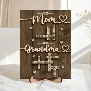 May include: Wooden sign with the words "First Mom, Now Grandma" in a cursive font. The sign includes names in a crossword puzzle style, with small heart accents. The sign is supported by a wooden stand.