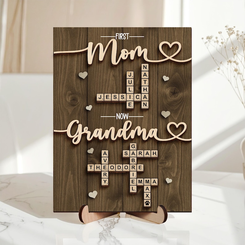 May include: Wooden sign with the words "First Mom, Now Grandma" in a cursive font. The sign includes names in a crossword puzzle style, with small heart accents. The sign is supported by a wooden stand.