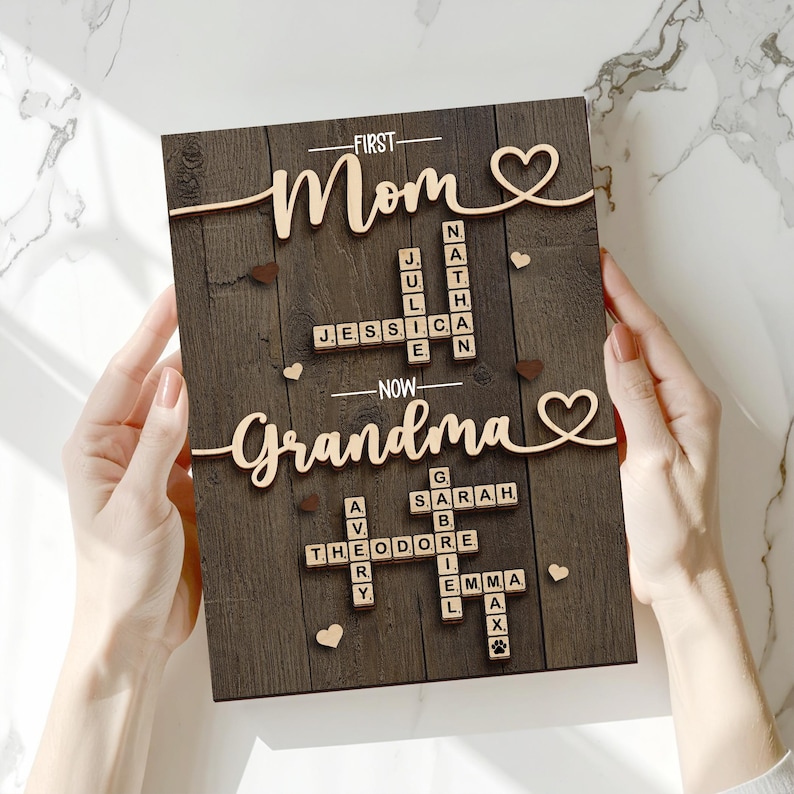 May include: Wooden sign with the words "First Mom Now Grandma" in a cursive font. The sign has a dark brown wood grain background with names spelled out in Scrabble-like tiles. Small wooden hearts and paw prints are scattered across the surface.