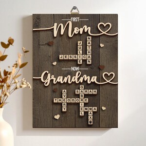 May include: Wooden wall art with the words "First Mom" and "Now Grandma" in cursive. The artwork features names spelled out in Scrabble-like tiles, with heart accents and a dark wood grain background. A metal hanger is at the top.
