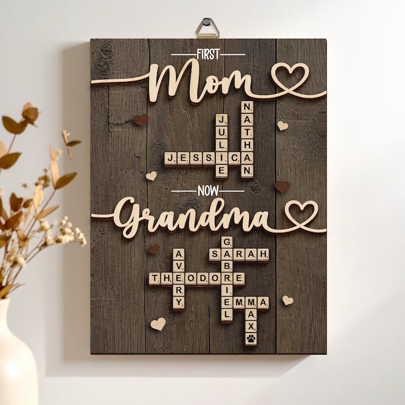 May include: Wooden wall art with the words "First Mom" and "Now Grandma" in cursive. The artwork features names spelled out in Scrabble-like tiles, with heart accents and a dark wood grain background. A metal hanger is at the top.