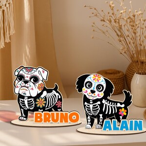 May include: Two wooden dog figurines with Day of the Dead-style designs. One is a bulldog with the name "BRUNO" in orange letters, and the other is a black dog with the name "ALAIN" in blue letters. Both have skeleton details.