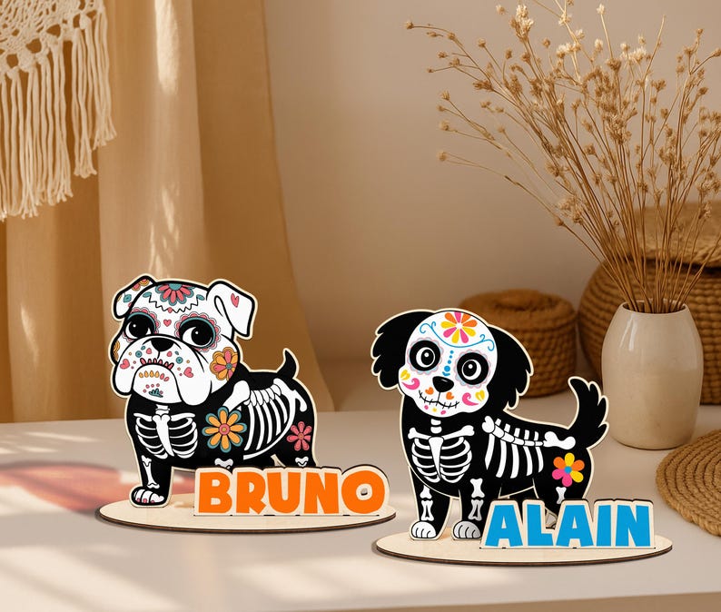 May include: Two wooden dog figurines with Day of the Dead-style designs. One is a bulldog with the name "BRUNO" in orange letters, and the other is a black dog with the name "ALAIN" in blue letters. Both have skeleton details.