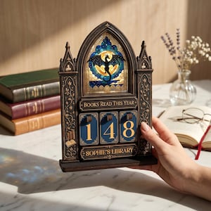 May include: A dark brown wooden book tracker with a gothic arch design, featuring a stained glass-style dragon insert, the text "BOOKS READ THIS YEAR" and "SOPHIE'S LIBRARY," and three rotating blue number tiles currently displaying the number 148.