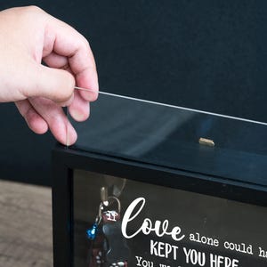 May include: A black shadow box with a clear glass front. The box contains keys and a decorative message in white script that reads "Love alone could have kept you here". A hand is shown lifting the glass front.