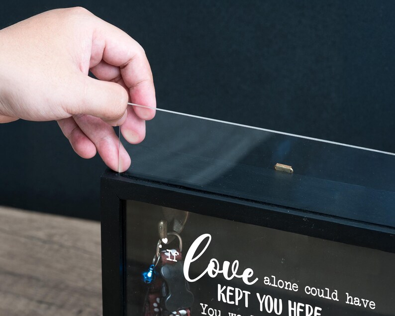 May include: A black shadow box with a clear glass front. The box contains keys and a decorative message in white script that reads "Love alone could have kept you here". A hand is shown lifting the glass front.