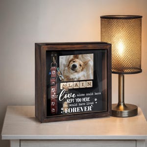 May include: A wooden shadow box with a glass front, displaying a photo of a dog, scrabble tiles spelling "ALAIN", and the text "If love alone could have kept you here, you would have lived forever." A dog collar and leash hang inside.