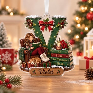 May include: A Christmas ornament shaped like the letter V, featuring a person reading a book in a chair with a cat, a stack of books labeled All I Want For Christmas Is Books, a red bow, and a wooden nameplate with the name Violet printed on it.