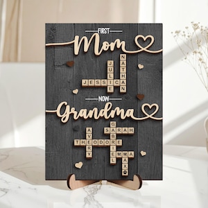 May include: A wooden sign with the words "First Mom" and "Now Grandma" in cursive script. The sign features a dark gray background with wooden accents, including hearts and names spelled out in Scrabble-like tiles. The sign is supported by a wooden stand.