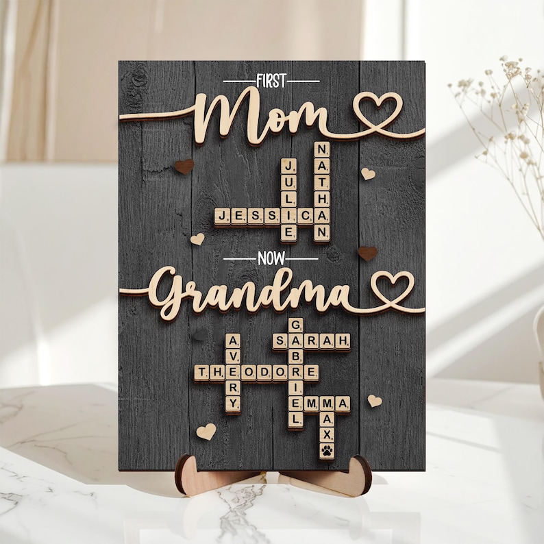 May include: A wooden sign with the words "First Mom" and "Now Grandma" in cursive script. The sign features a dark gray background with wooden accents, including hearts and names spelled out in Scrabble-like tiles. The sign is supported by a wooden stand.