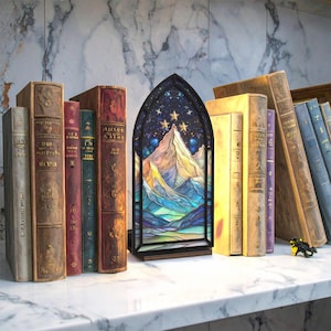 May include: A decorative stained glass lamp featuring a mountain scene under a starry night sky. The lamp is framed in black and sits on a wooden base. It is displayed among a collection of antique books with various colored spines.