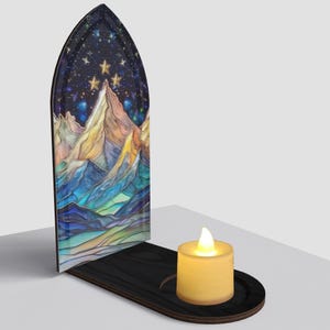 May include: A decorative candle holder featuring a mountain landscape design with a starry night sky. The holder has a black wooden base with a cutout for a battery-operated candle. The mountain scene is rendered in shades of blue, yellow, and purple.