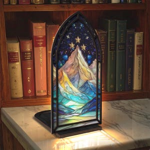 May include: A stained glass bookend featuring a mountain landscape under a starry night sky. The bookend has a black frame and a sturdy base. The artwork uses a palette of blues, greens, yellows, and purples. The bookend is displayed on a marble surface.