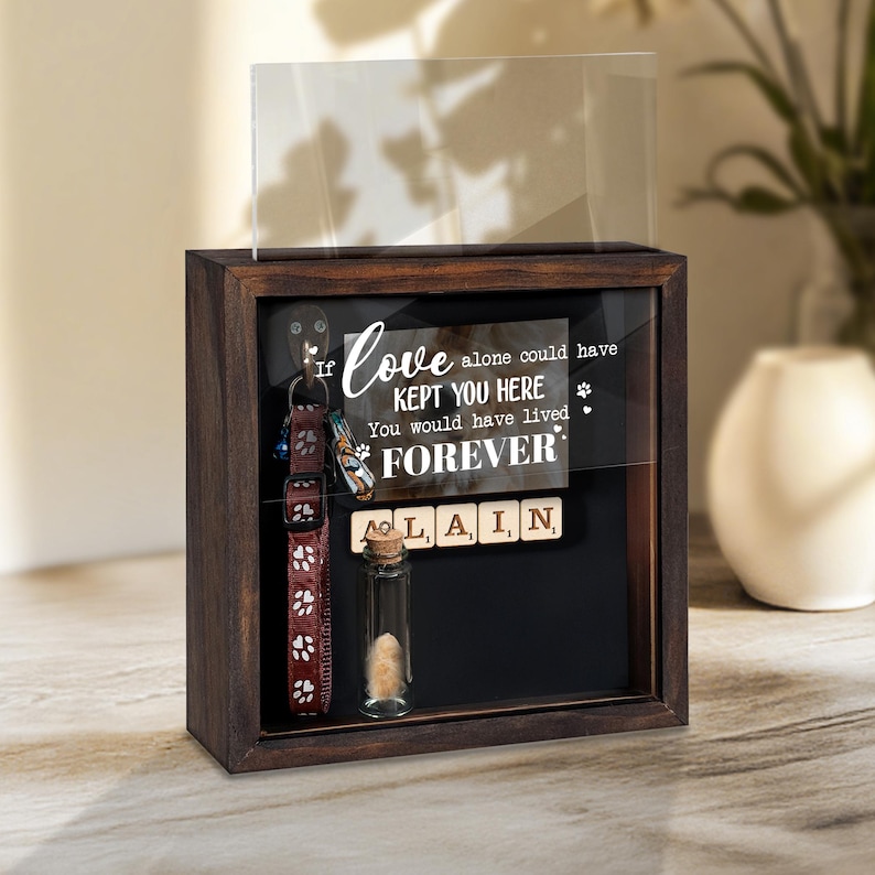 May include: A dark wooden shadow box with a clear glass front. Inside, a pet collar, a small glass vial, and a sign that reads "If love alone could have kept you here, you would have lived forever."
