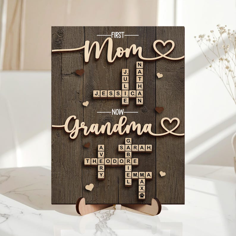 May include: Wooden sign with the words "First Mom" and "Now Grandma" in a cursive font. The sign has a dark wood grain background with names spelled out in a crossword puzzle style. Small heart and paw print accents are included.
