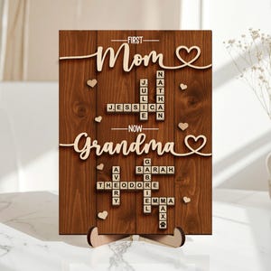 May include: Wooden plaque with the words "First Mom, Now Grandma" in a cursive font. The plaque features names spelled out in Scrabble tiles and heart-shaped accents. The background is a dark wood grain.