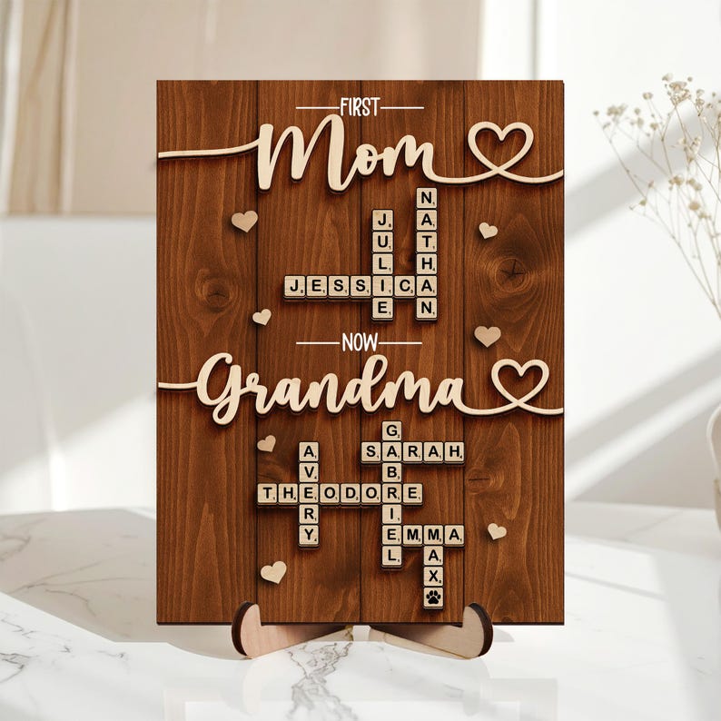 May include: Wooden plaque with the words "First Mom, Now Grandma" in a cursive font. The plaque features names spelled out in Scrabble tiles and heart-shaped accents. The background is a dark wood grain.