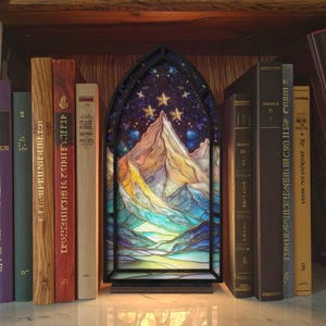 May include: A decorative stained glass panel depicting a mountain range under a starry sky, framed in black. The panel is illuminated and placed on a shelf with various books. The color palette includes blues, yellows, and purples.