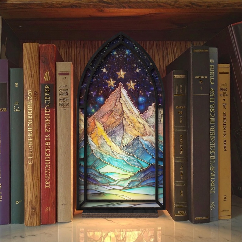 May include: A decorative stained glass panel depicting a mountain range under a starry sky, framed in black. The panel is illuminated and placed on a shelf with various books. The color palette includes blues, yellows, and purples.
