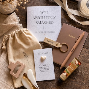May include: A gift set containing a greeting card with the text YOU ABSOLUTELY SMASHED IT, a brown notebook, a beige pen with BE KIND text, a beige keychain with the word Loved, a small card with a heart and HERE'S A HUG text, a tube of hand cream, and a hair clip.