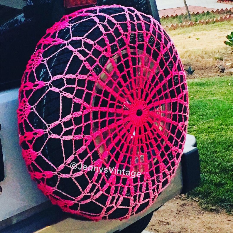 May include: A hot pink, handmade crochet spare tire cover. The cover is a web-like design, with a central circular pattern and radiating lines. The text "@JennysVintage" is visible on the cover.