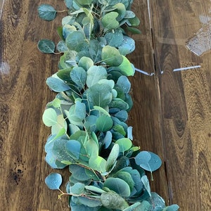 May include: A long garland of eucalyptus leaves, with a light green color, is laid out on a wooden surface. The leaves are arranged in a flowing pattern, creating a natural and elegant look.