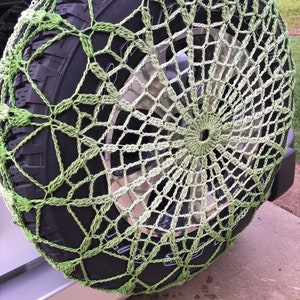 May include: A green and off-white crocheted spare tire cover. The cover is made of a net-like pattern and is stretched over a black tire. The tire is mounted on a vehicle.