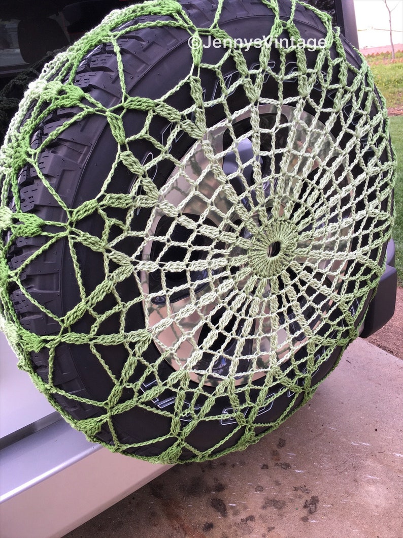 May include: A green and off-white crocheted spare tire cover. The cover is made of a net-like pattern and is stretched over a black tire. The tire is mounted on a vehicle.