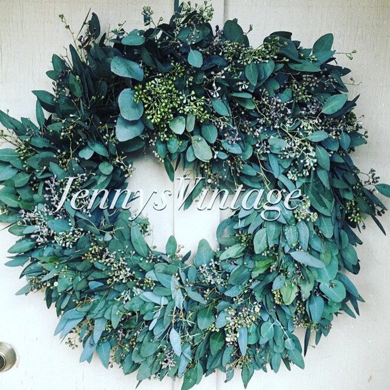 May include: A circular wreath made of green eucalyptus leaves and small white flowers. The wreath is displayed against a white background. The text "Jennys Vintage" is written across the wreath.