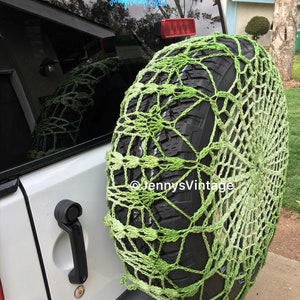 May include: A green crocheted tire cover on a spare tire. The cover is a net-like design with varying shades of green. The spare tire is mounted on the back of a white vehicle. The text "JennysVintage.etsy.com" is visible.