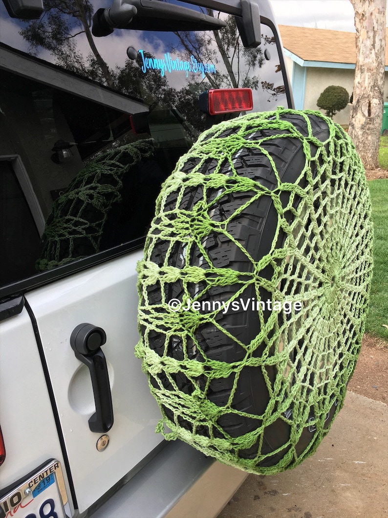 May include: A green crocheted tire cover on a spare tire. The cover is a net-like design with varying shades of green. The spare tire is mounted on the back of a white vehicle. The text "JennysVintage.etsy.com" is visible.