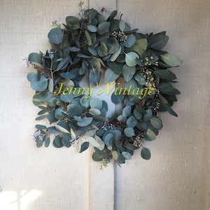 May include: A round eucalyptus wreath with green and blue-green leaves and small white flowers. The wreath is hanging on a light-colored wooden door. The text "Jenny's Vintage" is written across the bottom of the wreath.
