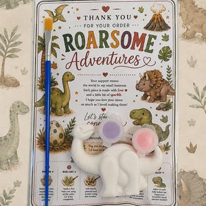 Puede incluir: Un kit de manualidades que incluye una figura de dinosaurio de yeso blanco, un pincel azul con cerdas amarillas y dos pequeños botes de pintura en morado y rosa. Los artículos vienen sobre una tarjeta con ilustraciones de dinosaurios y el texto Roarsome Adventures.
