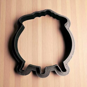 May include: A black plastic cauldron-shaped cookie cutter. The cutter has a rounded body with a wavy top edge and a curved base with three rounded feet. It is designed for making biscuits or other treats.