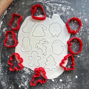 May include: A top-down view shows red Christmas-themed cookie cutters surrounding cut-out shapes in rolled-out dough. The cutters include a Santa hat, stocking, snowman, reindeer, gingerbread man, and Christmas tree.