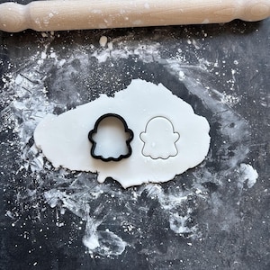 May include: A black ghost-shaped cookie cutter and a ghost-shaped cutout in white dough. A wooden rolling pin is above the dough on a dark surface dusted with flour. The image suggests baking or crafting activities.