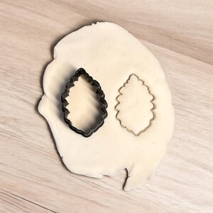 May include: A black metal cookie cutter in the shape of a leaf, with a leaf shape cut out of white fondant.