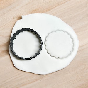 May include: Two round cookie cutters on a white surface. The cutter on the left is black and the cutter on the right is silver.