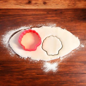May include: A red plastic seashell cookie cutter on a white surface with a cutout of the seashell shape. The surface is covered in white powder.