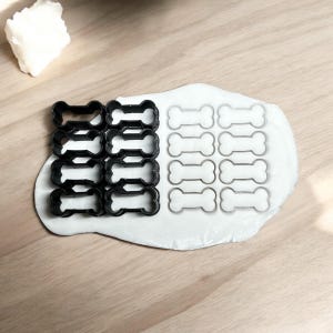 May include: A set of 12 bone-shaped cookie cutters in black and white. The cutters are arranged in two rows of six, with the black cutters on the left and the white cutters on the right. The cutters are on a white surface.