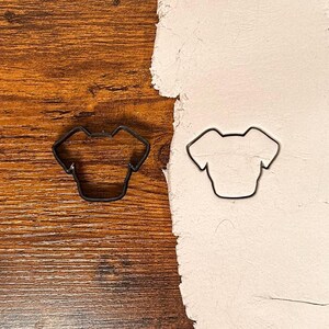 May include: Two black metal cookie cutters in the shape of a t-shirt. One cutter is on a wooden surface, and the other is on a white surface.