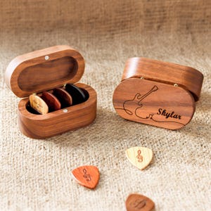 Puede incluir: Una caja de madera con una guitarra y el nombre "Skylar" grabado en la tapa. La caja está abierta y contiene un juego de púas de guitarra. Las púas son de madera y tienen diferentes colores y diseños.