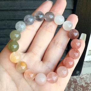 May include: A beaded bracelet featuring a variety of colorful, translucent gemstone beads. The beads range in color from clear to shades of green, yellow, orange, pink, and brown. The bracelet is displayed on a hand, showcasing the natural variations in the stones.