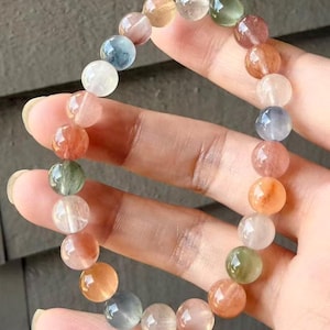 Puede incluir: Un brazalete de cuentas con una variedad de piedras preciosas redondas y coloridas. Las cuentas son de color naranja, rosa, verde, azul y transparente. El brazalete está ensartado en una banda elástica y lo sostiene una mano.