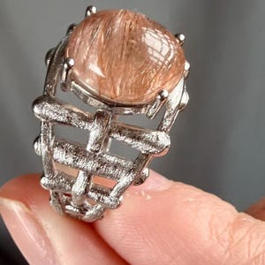 May include: A silver ring with a large, round, translucent gemstone with reddish-brown inclusions. The ring has a textured, woven-like design around the gemstone, creating a unique piece of jewellery.