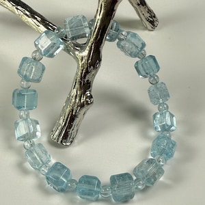May include: A bracelet composed of light blue, translucent, cube-shaped beads alternating with small, clear, round spacer beads. The bracelet is draped over a silver-colored, branch-shaped display stand with a smooth, metallic finish.