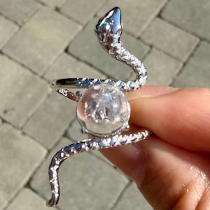 May include: A silver-toned ring shaped like a coiled snake with a textured scale pattern along its body. The snake wraps around a central, clear, spherical stone that contains visible internal inclusions. The metal band is stamped with S925.
