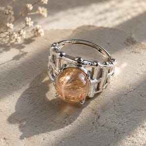 May include: A silver-toned ring featuring a round, translucent rutilated quartz gemstone with visible golden needle-like inclusions. The band has a textured, cage-like design with vertical bars and small spherical accents surrounding the central stone.