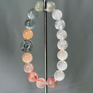 Peut inclure: Un bracelet composé de perles rondes translucides présentant un dégradé de couleurs allant du transparent et vert pâle au pêche, orange et rose rougeâtre. Une breloque circulaire en métal argenté avec une pierre bleue centrale est intégrée.