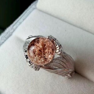 May include: A silver-toned ring featuring a central, circular, translucent stone with copper-colored, fibrous inclusions. The band is designed with a textured, leaf-like motif on both sides, accented by small, clear, sparkling stones embedded along the metal.